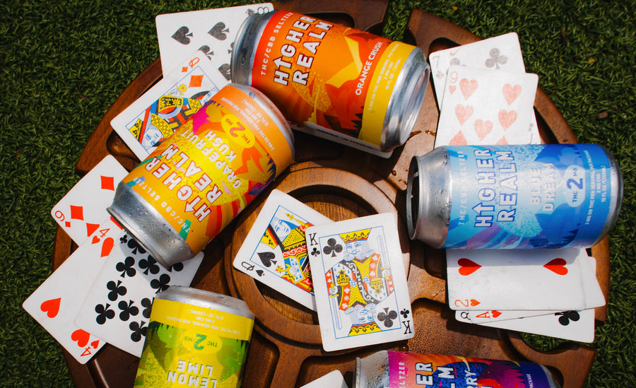 Three cans of Higher Realm THC drink are arranged on a wooden tray with playing cards on a green grass-like surface. The visible cans include yellow, orange, and blue flavors, with the center can reading Higher Realm Blue Dream