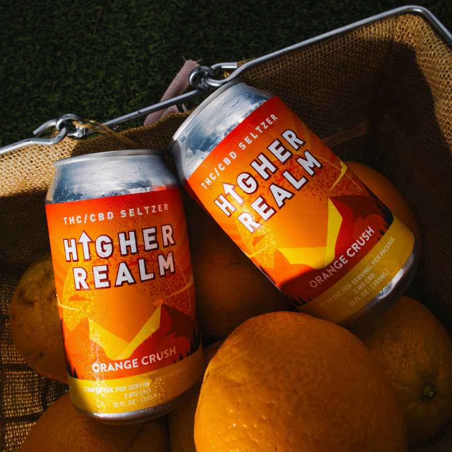 Two Higher Realm Orange Crush THC and CBD drinks in a tweed-lined basket surrounded by fresh oranges.